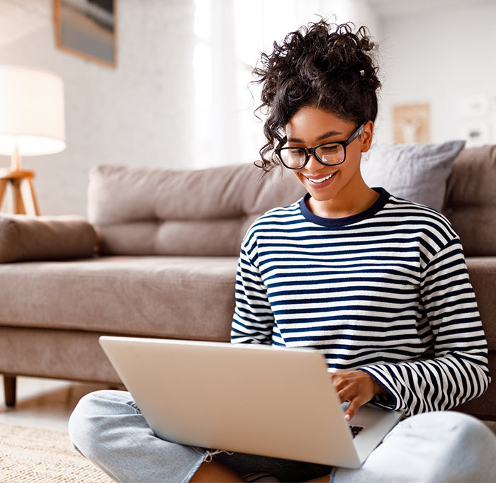 Woman in striped shirt sitting in front of couch and typing on laptop