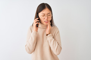 Woman with glasses holding phone and rubbing jaw in pain